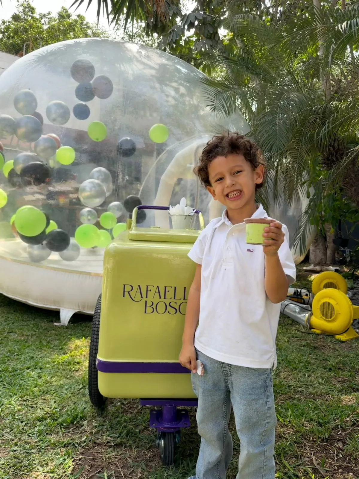 Niño sonriendo con un pote de gelato de chocolate frente al carrito heladero, en una fiesta con un inflable y globos de fondo