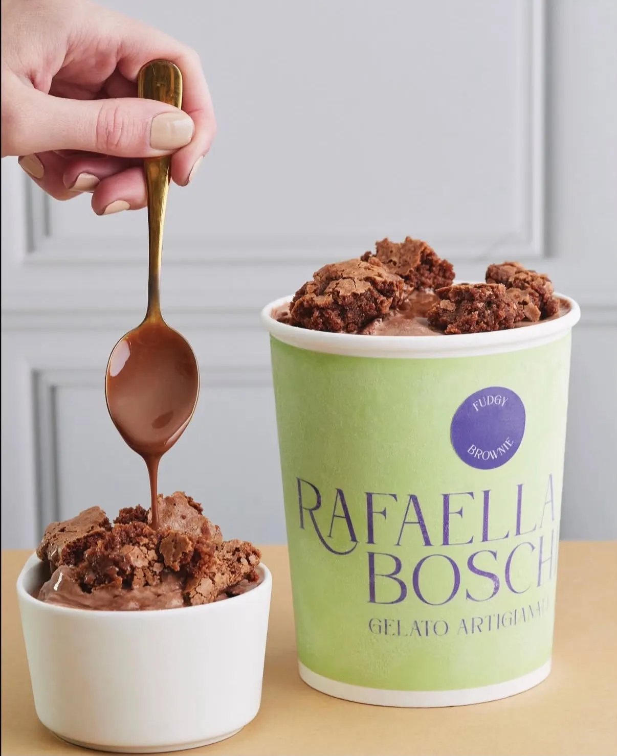 Pote de gelato de chocolate cubierto con trozos de brownie, con una cuchara sirviendo caramelo sobre un bowl del mismo gelato