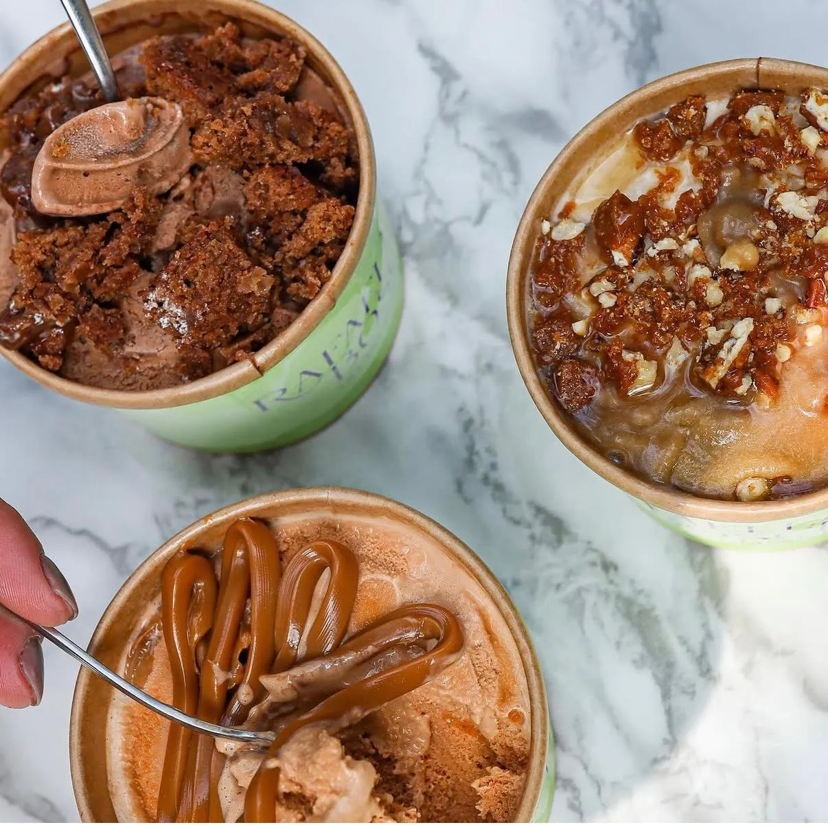 Tres potes de gelato sobre mármol: brownie de chocolate, caramelo con pecanas y dulce de leche